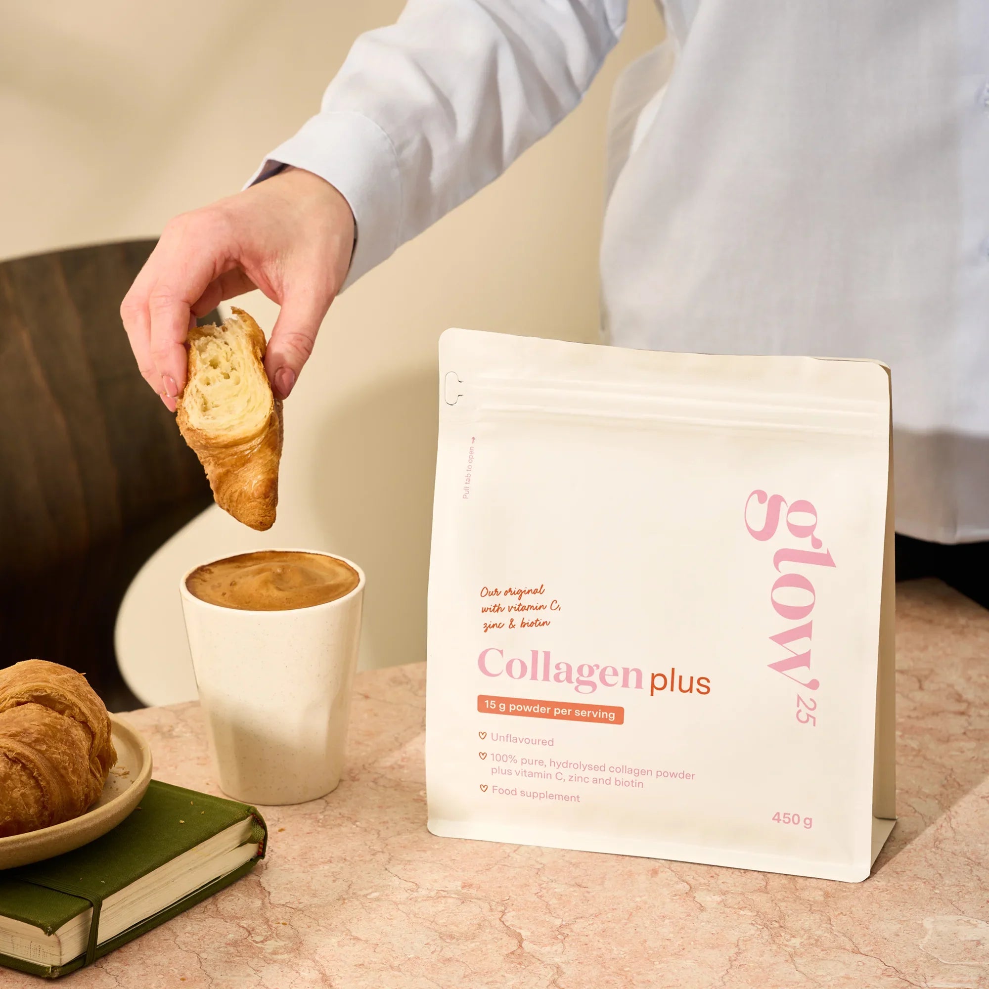 Hand dipt croissant in een kopje koffie met Glow25 Collageen Plus tijdens ontbijt, eenvoudig in gebruik.