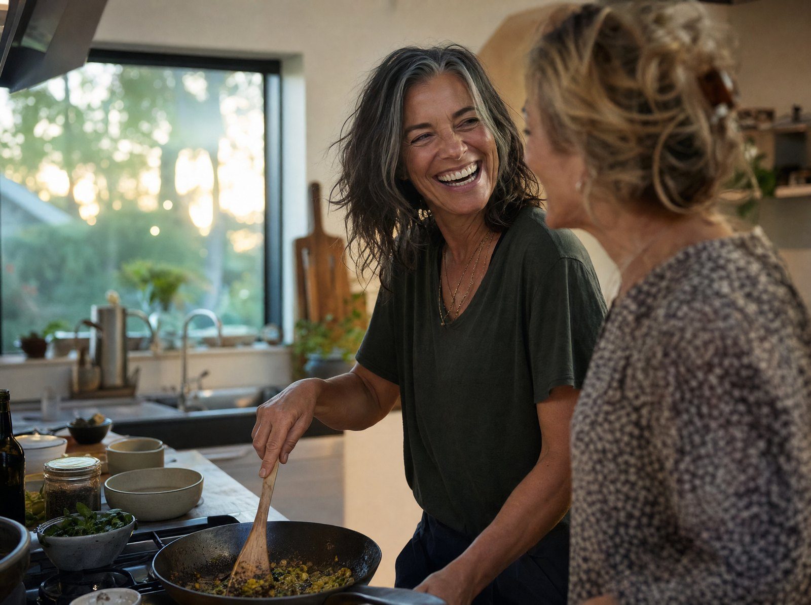 Donna intorno ai 50 anni ride la sera cucinando con un'amica, piena di energia
