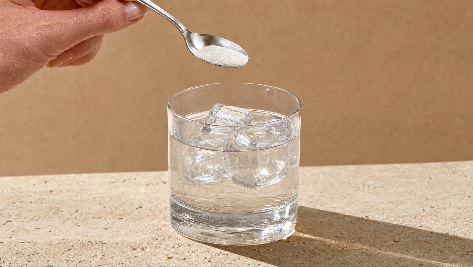 Un cucchiaino versa una polvere bianca in un bicchiere d'acqua con cubetti di ghiaccio